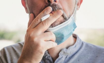 A close up of a man wearing a disposable mask over his chin, holding a cigarette to his lips. 